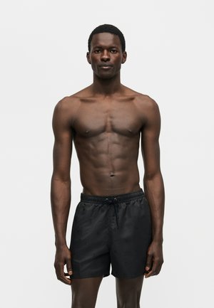 Μυώδης άνδρας με σκουρόχρωμο δέρμα στέκεται αντικρίζοντας μπροστά, φοράει μαύρο μαγιό με κορδόνι σε απλό λευκό φόντο.