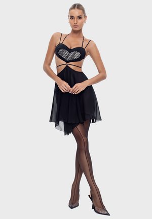 Woman wearing a black strappy dress with a heart detail, sheer black stockings with a back seam, and black high-heeled shoes, posing.