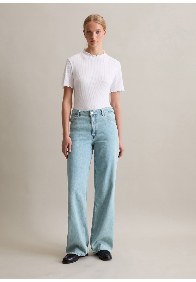 Weißes, geripptes Oberteil mit gewellten Kanten, kombiniert mit hellblauen, weit geschnittenen Jeans. Glatte Textur und klassisches Design mit fünf Taschen. Schwarze Schuhe.