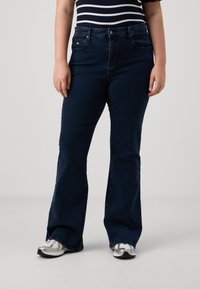 Mørkeblå utsvingte jeans laget av denim, med en frontlomme og beltehemper. Brukt sammen med en stripete topp og sølvfargede joggesko.
