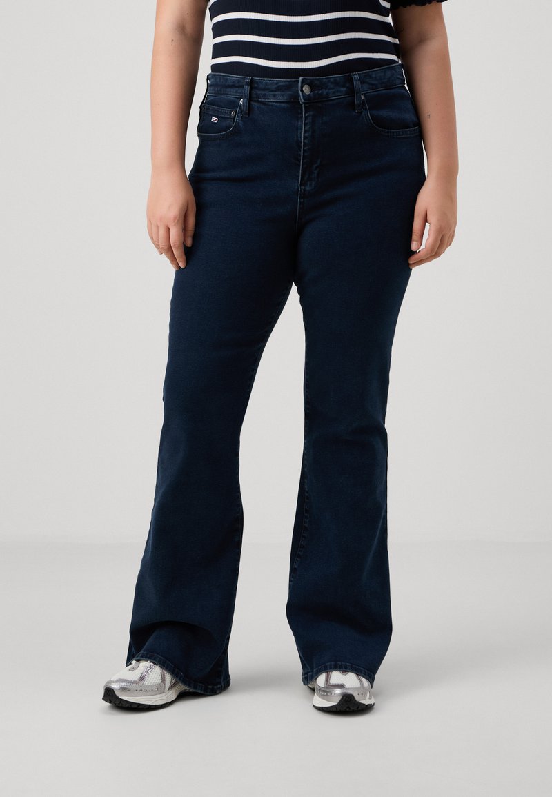 Jeans de mezclilla azul oscuro con campana, que cuentan con un bolsillo frontal y presillas para cinturón. Llevados con una blusa a rayas y zapatillas plateadas.