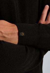 Zwarte gebreide trui met een gestructureerd patroon, geribbelde mouwen en een klein merkembleem op de mouw. Zacht materiaal met een comfortabele pasvorm.