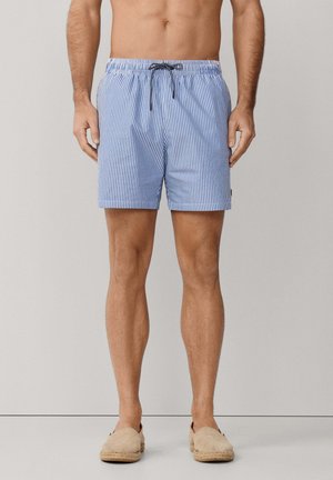 Man met blauw-wit gestreepte korte broek met verticale strepen, zwarte trekkoord en beige instapschoenen, staand tegen een effen lichte achtergrond.