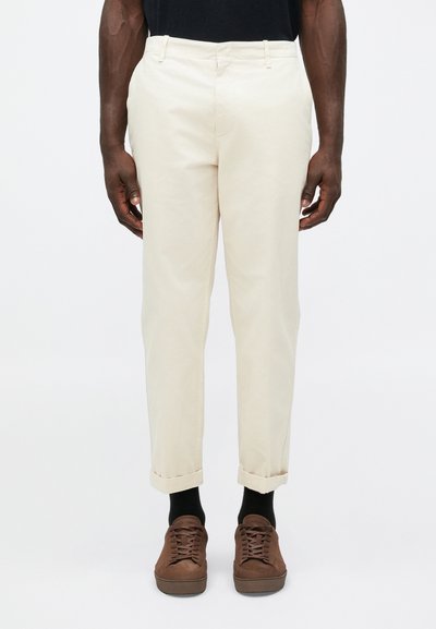 Beige cotton chinos with a straight fit, rolled cuffs, and front pockets. Worn with brown sneakers and black socks.