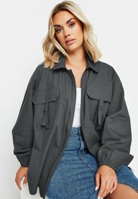 Veste zippée grise avec un col, deux poches avant et des poignets élastiques ; portée sur un haut blanc et une jupe en denim. Texture lisse.