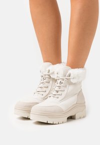 Botas blancas de invierno con suelas gruesas, cordones en la parte delantera y cuellos de piel esponjosa, llevadas sobre piernas desnudas frente a un fondo liso.