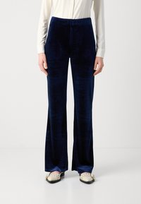 Námornícke modré zamatové plisované nohavice s hladkou textúrou, vysokým pásom a bočnými vreckami. Styled s krémovou košeľou na gombíky a svetlými topánkami.