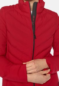 Chaqueta acolchada roja con cuello alto, con costuras en forma de chevron, cremallera negra y puños ajustables. Textura suave y diseño ligero.