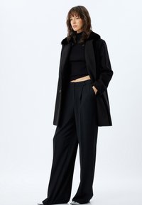 Cappotto di lana nero con collo in pelliccia sintetica, abbinato a un top corto nero a coste e pantaloni neri a gamba larga, con una silhouette rilassata.