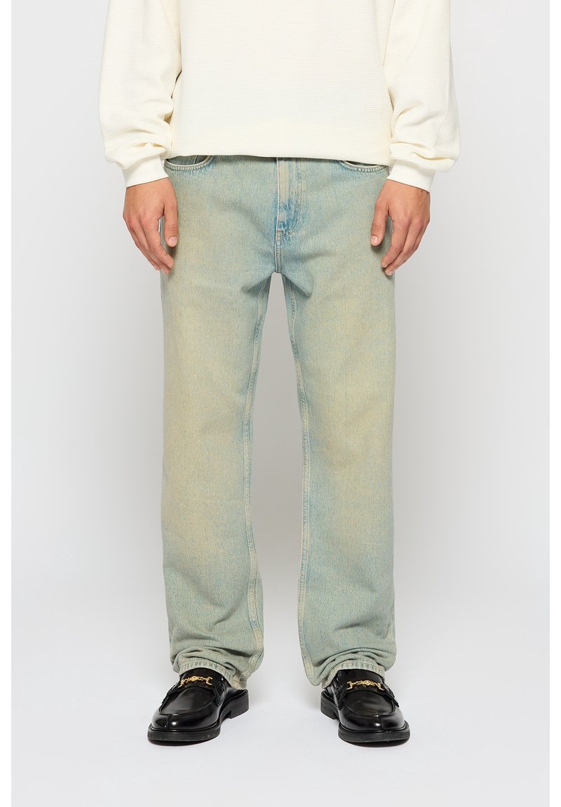 Homme portant un jean bleu clair délavé, un pull crème à manches longues et des mocassins noirs en cuir avec un détail chaîne dorée, debout sur fond blanc.