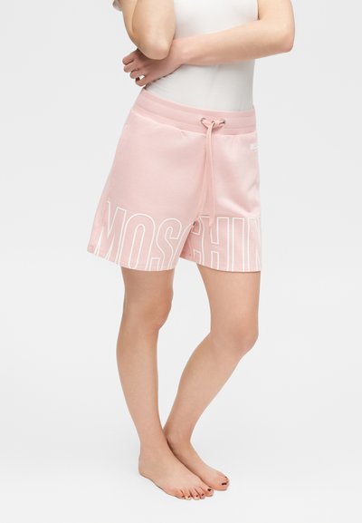 Άτομο που φοράει ανοιχτό ροζ σορτς με κορδόνι και μεγάλο κείμενο "MOSCHINO" στα πόδια και λευκή αμάνικη μπλούζα, στέκεται ξυπόλητο.