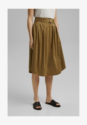 Femme portant une jupe midi marron taille haute avec des plis et une ceinture, associée à des sandales plates noires et un haut blanc rentré.