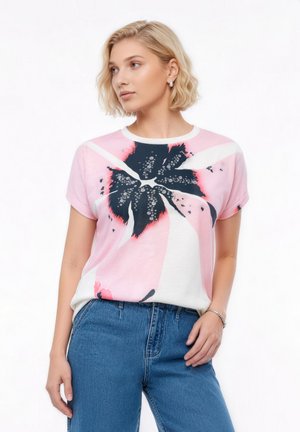 Vrouw met kort blond haar, gekleed in een roze-wit T-shirt met een groot zwart bloemenmotief en blauwe spijkerbroek, staand tegen een witte achtergrond.