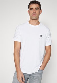 T-shirt bianco in cotone con collo rotondo, maniche corte e logo navy sul lato sinistro del petto. Il tessuto appare liscio e leggero.