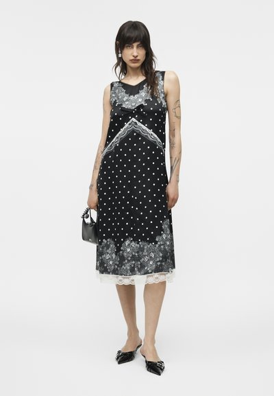 Γυναίκα με σκούρα μαλλιά, φορώντας μαύρο μίντι φόρεμα χωρίς μανίκια με λευκές πουά, λουλουδένια δαντελένια σχέδια και κρατώντας μια μικρή μαύρη τσάντα χειρός.