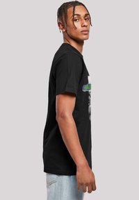 Melna kokvilnas T-krekls ar īsām piedurknēm, kas ietver grafisko dizainu un tekstu zaļā un baltā krāsā. Klasisks piegriezums ar apļa izgriezumu.