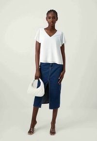 Topbianco a maniche corte con scollatura a V, abbinato a una gonna di jeans con spacco frontale. Tiene in mano una borsa bianca testurizzata e indossa sandali marroni.