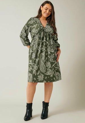 Robe verte à motif paisley avec un décolleté en V, des manches longues et une taille froncée. Associée à des bottines noires, avec une texture lisse.
