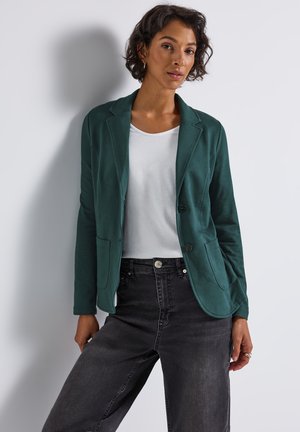 Blazer teal avec une coupe ajustée, deux poches avant et fermeture à un bouton, porté sur un t-shirt blanc et un jean noir taille haute.