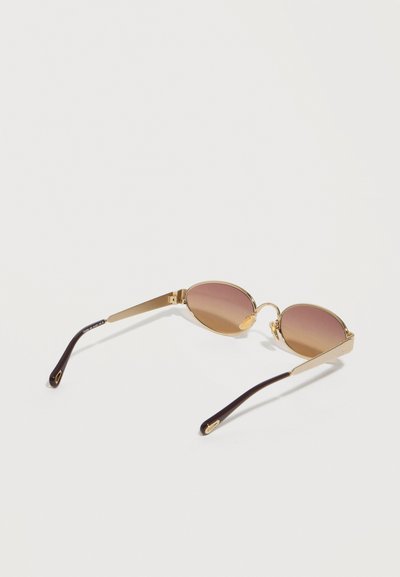 Οβάλ γυαλιά ηλίου με χρυστά μεταλλικά πλαίσια και καφέ φακούς με διαβάθμιση, ακουμπισμένα σε λευκή επιφάνεια.