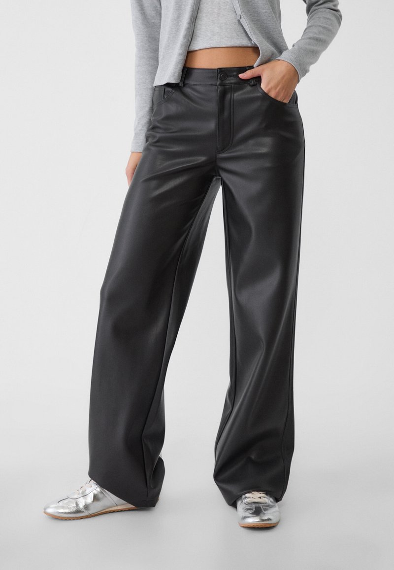 Personne portant un pantalon en cuir noir taille haute à jambes larges, un top gris court, un cardigan gris et des chaussures argentées à lacets, debout la main dans la poche.