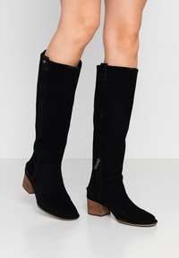Black suede knee-high boots with a pointed toe, zip closure on the side, and a stacked wooden heel. Smooth texture and sleek design.