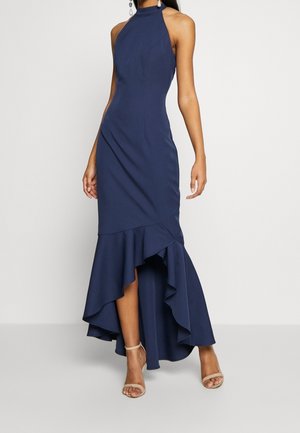 Mujer con un vestido azul marino sin mangas de largo irregular con dobladillo con volantes y zapatos de tacón beige con tira en el tobillo sobre un fondo sencillo.