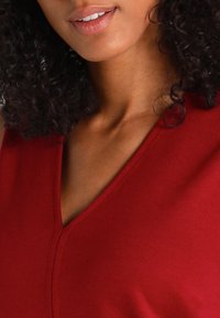 Červený top bez rukávů s výstřihem do V, hladkou texturou a jemnými švy podél výstřihu a ramene.