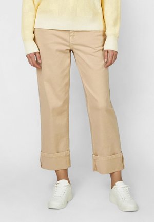 Person trägt beige kurze Hosen mit breiten umgeschlagenen Bündchen, weiße Turnschuhe und einen gelben Pullover und steht vor einem einfarbigen Hintergrund.