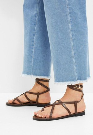 Sandali flat in pelle marrone con cinturini intrecciati, fibbia alla caviglia regolabile e design minimalista, abbinati a jeans cropped azzurri.