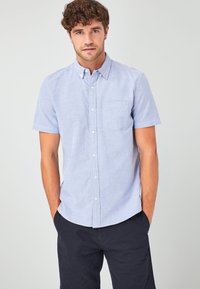 Camisa de manga corta, con cuello, en tejido texturizado azul claro, con un bolsillo frontal y botones blancos, combinada con pantalones cortos oscuros.