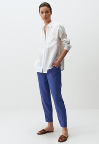 Witte linnen overhemd met een kraag en een voorzak, gedragen met blauwe tapered broeken en bruine strappy sandalen op een neutrale achtergrond.