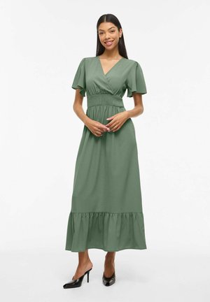 Kobieta z długimi ciemnymi włosami, ubrana w zieloną sukienkę kopertową z falbanowymi rękawami i czarne szpilki, stojąca i uśmiechająca się na białym tle.