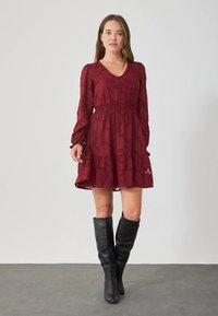 Γυναίκα που φοράει ένα μακρυμάνικο μπορντώ φορεμα με λουλουδάτη υφή και μαύρες μπότες μέχρι το γόνατο, μπροστά σε απλό ανοιχτό φόντο.