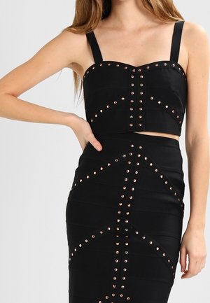 Femme portant un crop top noir sans manches et une jupe taille haute avec des motifs de clous en cuivre, posant avec une main sur la hanche.