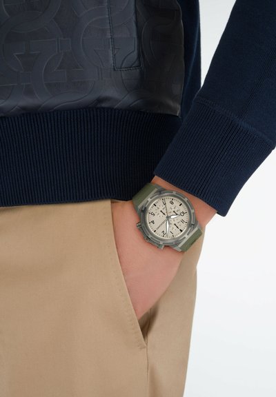 Herrenarmbanduhr mit grauer Textur, weißem Zifferblatt, schwarzen Markierungen und grünem Silikonarmband. Am Handgelenk getragen, kombiniert mit einem marineblauen Pullover und beigen Hosen.