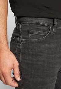 Vaqueros de mezclilla gris oscuro con un corte ajustado, con bolsillos estándar, cierre de botones y detalles sutiles de costura. Mano apoyada al lado.