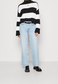 Pull en maille à rayures noires et blanches avec un col cheminée, associé à un jean large bleu clair et des bottes noires épaisses.