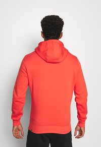 Uomo con capelli ricci che indossa un luminoso hoodie rosso, girato di spalle su uno sfondo bianco semplice.