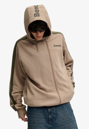 Person iført beige Bench zip-up hættetrøje med hætten oppe og mørke solbriller, kombineret med blå jeans, stående mod en ensfarvet baggrund.