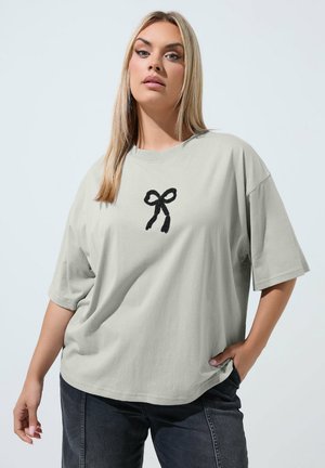 Blondine vrouw die een losse lichtgrijze T-shirt draagt met een klein zwart strikje op de borst en donkere jeans, staand met één hand in de zak.