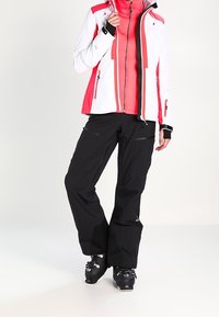 Giacca da sci bianca con accenti rosa e neri; chiusura frontale con zip, collo alto. Pantaloni neri, vestibilità ampia, aree rinforzate, abbinati a scarponi da sci.