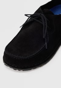 Schwarze Wildleder-Schnürschuhe mit genähten Details und strukturiertem Sohlen auf einem hellen Hintergrund, der Front- und Seitenansichten zeigt.