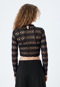 Sort blonde crop top med lange ærmer, med en bølget kant og nøglehul detalje bagpå. Tekstureret med vandrette striber af blonde.