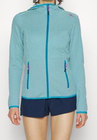 Giacca con cappuccio e zip di colore blu chiaro con un motivo esagonale. Presenta due tasche frontali con zip e accenti di blu scuro in contrasto.