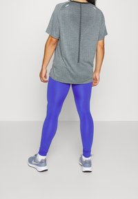 Top grigio a maniche corte con motivo testurizzato, dettaglio della cucitura posteriore, e leggings blu, abbinati a scarpe da ginnastica grigie.