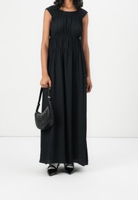 Black pleated maxi dress with cap sleeves, fitted bodice, and gathered waist. Accented with a studded black handbag and black pointed shoes.