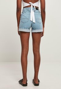 Pantalones cortos de mezclilla en azul claro, con dobladillos enrollados y dos bolsillos traseros. La cinturilla presenta un detalle de lazo blanco. Se llevan con zapatos planos negros.