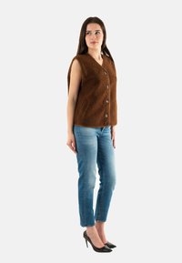 Gilet marrone senza maniche con scollatura a V e cinque grandi bottoni; realizzato in materiale morbido e testurizzato. Abbinato a jeans blu e tacchi neri.