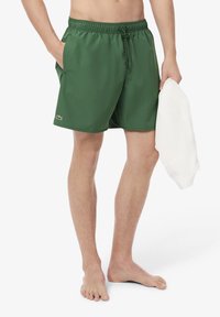 Shorts de bain verts avec taille élastique, poches latérales et petit logo Lacoste. Le tissu semble lisse et léger.